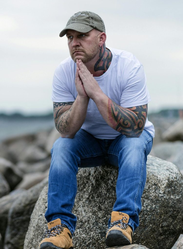 Contemplative man with tattoos sitting on a rocky shoreline, deep in thought.