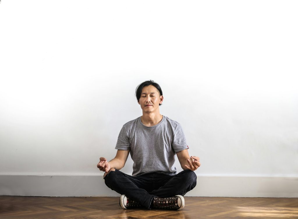 Asian man practicing meditation indoors on a wooden floor, promoting relaxation and mindfulness.