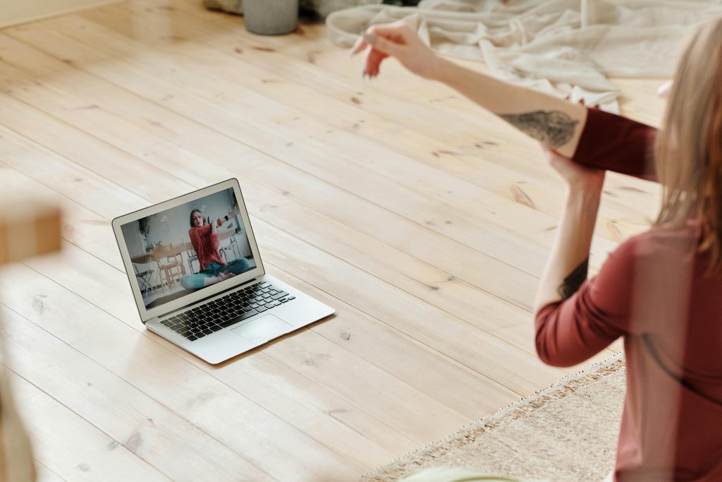 A woman stretches while following an online yoga class on a laptop in a cozy indoor setting.