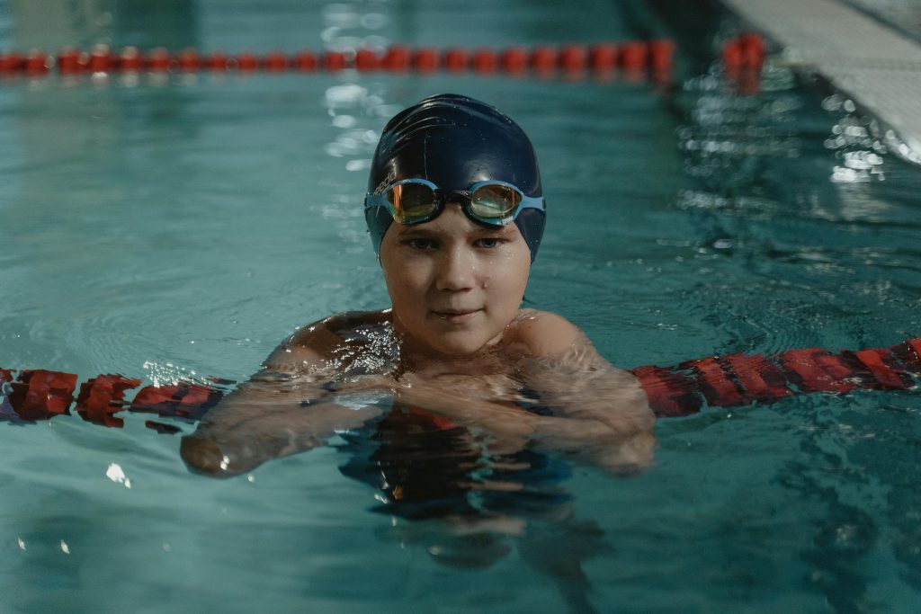 A young swimmer in a cap and goggles taking a break in an indoor pool.