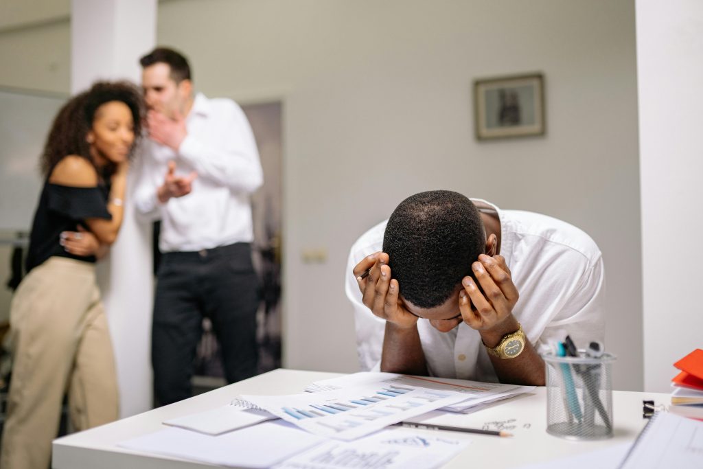 Despairing worker at office desk as colleagues gossip in background illustrating workplace bullying.